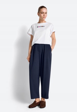 Kobieta stojąca, ubrana w biały T-shirt z wielokolorowymi uśmiechniętymi buźkami, granatowe spodnie o szerokich nogawkach oraz brązowe mokasyny, na tle jasnego, jednolitego tła.