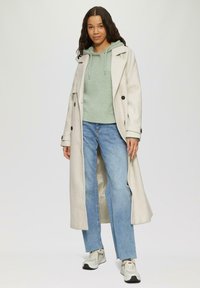 Langer beiger Mantel mit schwarzen Knöpfen über einem grünen Hoodie, kombiniert mit hellblauen Jeans und grauen Sneakers. Glattes Material und bequeme Passform.