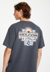 Musta harmaa puuvillapaita, jossa on värikäs graafinen kuva takana, jossa on aurinko, silmä ja timantti-muotoja. Paidassa on teksti "VOLCOM STONE 1991."
