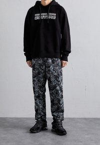 Černá mikina s kapucí s bílým logem, kombinovaná s černými kalhotami s propracovanými šedými vzory a grafickými motivy. Černé tenisky.