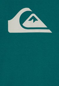 Teal bomull t-skjorte med et stort hvitt logo av en bølge og et fjell, glatt tekstur og korte ermer.