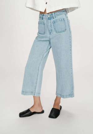 Jean large et court bleu clair porté avec des chaussures noires à enfiler à bout carré et une veste courte blanche, debout sur un fond uni.