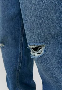 Blå denimjeans med slitna hål vid knäna, med en texturerad tyg och lätt blekta detaljer längs sömmarna.