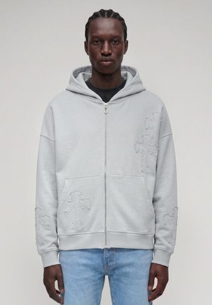 Man with braided hair wearing light gray zip-up hoodie with cross patterns and light blue jeans, standing against plain background.