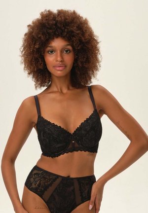 Schwarzer Spitzen-BH mit Unterbruststütze und Blumenmuster, ausgestattet mit verstellbaren Trägern und dekorativen gewellten Kanten. Passende durchsichtige, hochgeschnittene Panties.