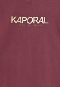 Tyrkysové tričko s metalickým zlatým nápisem "KAPORAL" a menším nápisem "letní slunce" pod ním v světle šedé barvě, vyrobené z bavlny.