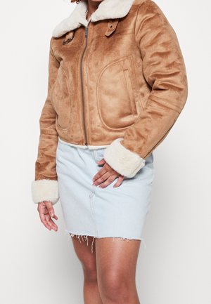 Frau trägt eine kurze beige Lammfelljacke mit weißem Fleece-Kragen und -Ärmeln, kombiniert mit einem hellblauen Jeans-Minirock mit ausgefranstem Saum.