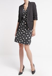 Zwart blazer over een bloemenpatroon jurk. De jurk heeft witte bloemen op een donkere achtergrond. Model draagt zwarte hoge hakken.