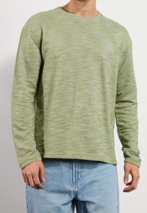 Homme portant un pull en tricot à manches longues vert clair et un jean bleu clair, debout devant un fond uni.