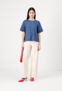 Top in denim a maniche corte con righe bianche verticali, abbinato a pantaloni larghi color crema e accessori rossi tra cui una cintura e décolleté a punta.