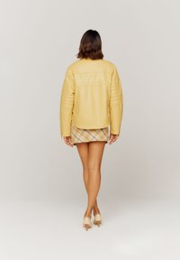 Gul lærjakke med quiltet design og bakbroderi som sier "TAKE A TRIP", kombinert med et rutete miniskjørt og nøytrale hæler.