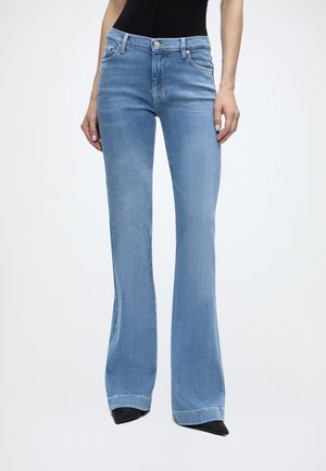 Person iført lyseblå højtaljede jeans med svaj og sort top, står med armene afslappet ned langs siderne mod en ensfarvet baggrund.
