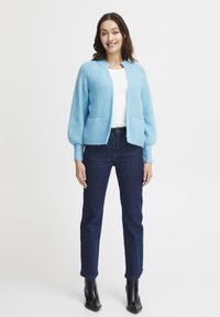 Cardigan a maglia blu chiaro con texture a coste, apertura frontale e due tasche frontali. Indossato sopra una maglietta bianca e abbinato a jeans di denim scuri e stivali neri.
