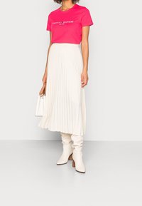 T-shirt in cotone rosa con logo "TOMMY HILFIGER", abbinato a una gonna midi bianca a pieghe e stivali al ginocchio color crema con tacco in legno.