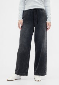 Črne široke kavbojke iz jeansa z elastičnim pasom in vrvico za zavezovanje. Imajo vidne šive in gladko teksturo. Združene z belimi supergami.