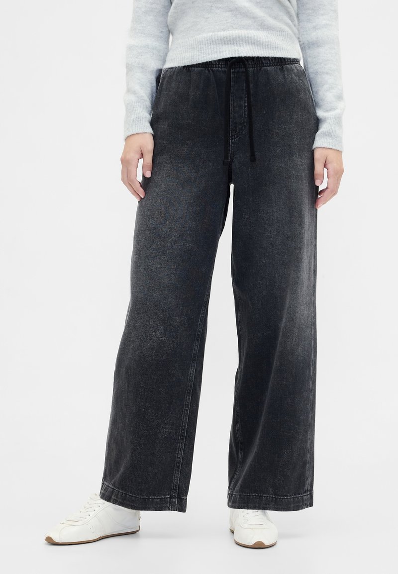 Črne široke kavbojke iz jeansa z elastičnim pasom in vrvico za zavezovanje. Imajo vidne šive in gladko teksturo. Združene z belimi supergami.