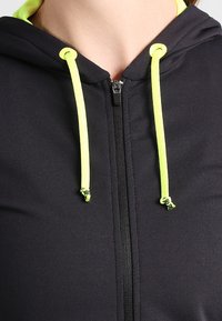 Gros plan sur un sweat à capuche noir zippé avec des cordons de serrage et des œillets jaune fluo autour de la zone du cou.