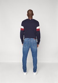 Navy modrý žebrovaný svetr s bílými a červenými pruhy na rukávech, v kombinaci se světle modrými džíny a bílými teniskami. Zaměřte se na strukturu a střih.