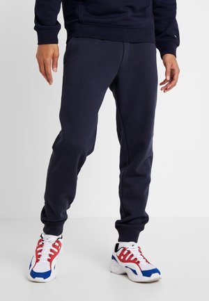 Unterkörper einer Person, die dunkelblaue Jogginghosen und weiße Sneakers mit roten und blauen Akzenten trägt, auf einem weißen Boden stehend.