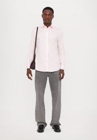 Camicia a maniche lunghe color rosa chiaro con bottoni, abbinata a jeans grigi a gamba larga e scarpe nere. Una piccola borsa scura è appesa a una spalla.