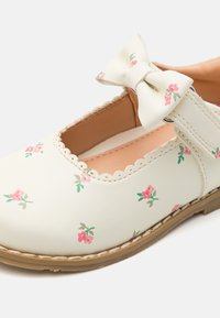 Kermapolttonen Mary Jane -kenkä, joka on valmistettu synteettisestä nahasta. Kengässä on kukkakuosi ja koristeelliset nauhat. Sirot reunat ja beige pohja.