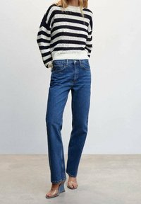 Gestrickter, gestreifter Pullover in Marineblau und Creme, kombiniert mit hoch taillierten blauen Denim-Jeans. Transparente Sandalen mit Absatz vervollständigen das Outfit.