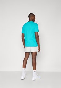 T-shirt turchese a maniche corte con scollatura a girocollo, abbinata a pantaloni corti beige texturizzati e sneaker bianche, vista da dietro.