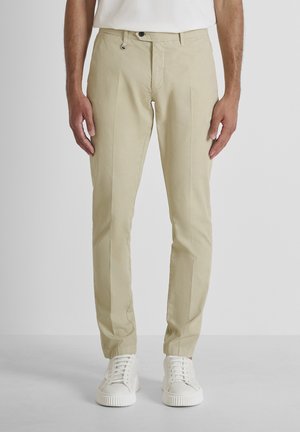 Homme portant un pantalon beige ajusté et des baskets blanches, debout les mains détendues le long du corps devant un fond uni.