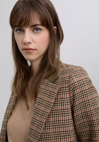 Blazer de pata de gallo marrón y crema con un cuello verde, con una tela texturizada y un diseño de apertura frontal, sobrepuesto a un suéter beige.