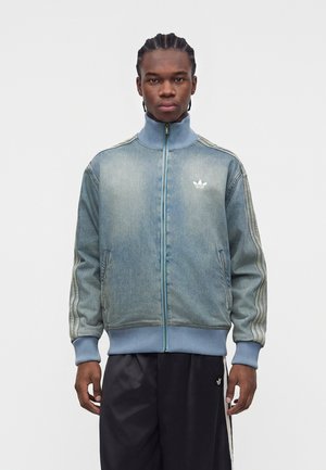 Joven con el pelo trenzado que lleva una chaqueta deportiva azul desgastada con cremallera y pantalones negros con rayas laterales sobre un fondo liso.