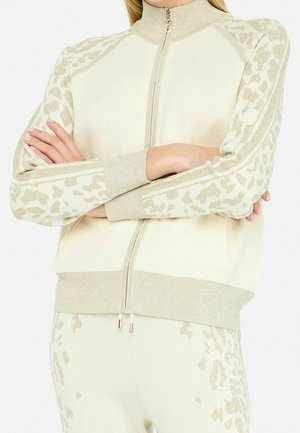 Personne portant une veste crème zippée et un pantalon avec des détails d’imprimé animal en or clair brillant, les bras croisés, visible des épaules aux hanches.