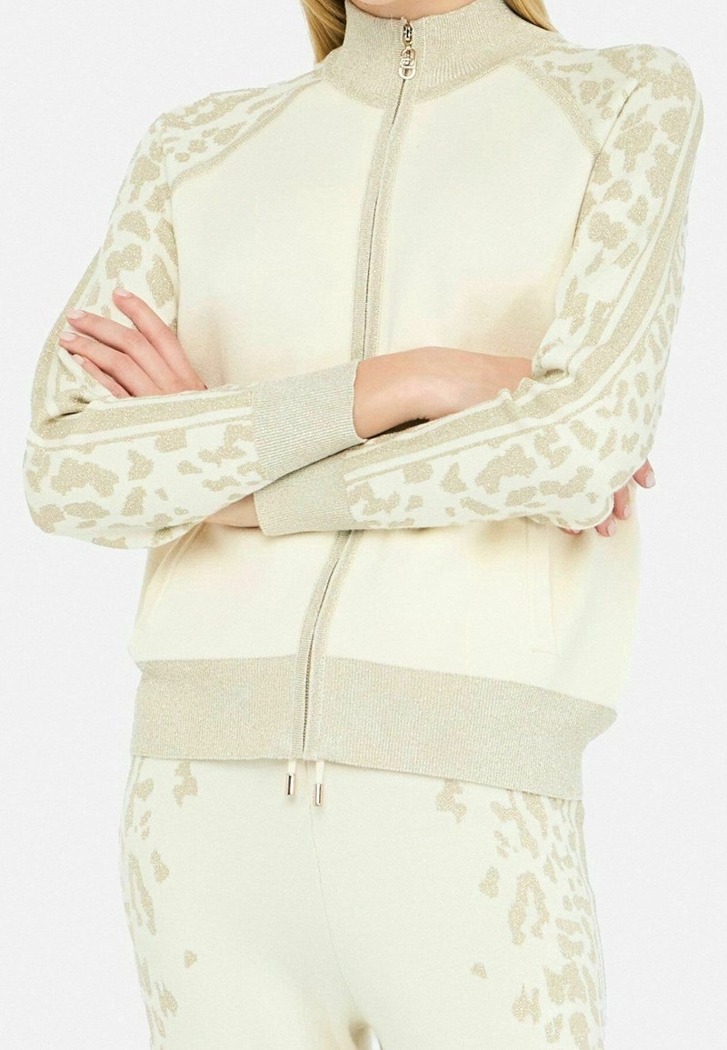 Personne portant une veste crème zippée et un pantalon avec des détails d’imprimé animal en or clair brillant, les bras croisés, visible des épaules aux hanches.