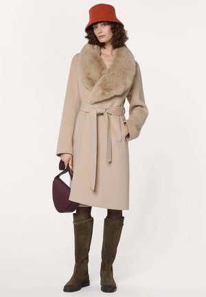 Vrouw draagt een beige jas met ceintuur en een grote bontkraag, een roestkleurige vissershoed, olijfgroene laarzen tot over de knie en houdt een donkerpaarse handtas vast.