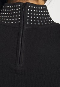 Svart stickad topp med hög krage prydd med silverfärgade nitar och en kort dragkedja på baksidan. Slät textur genom hela plagget.