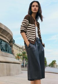Brązowo-kremowy sweter na guziki w paski, zestawiony z czarnymi, skórzanymi szortami z wysokim stanem. Modelka stoi z rękami w kieszeniach.