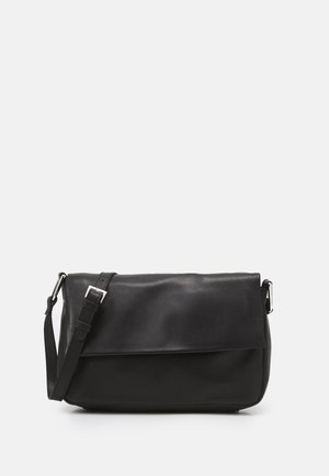 Sac bandoulière en cuir noir avec sangle ajustable et boucle argentée sur fond clair.