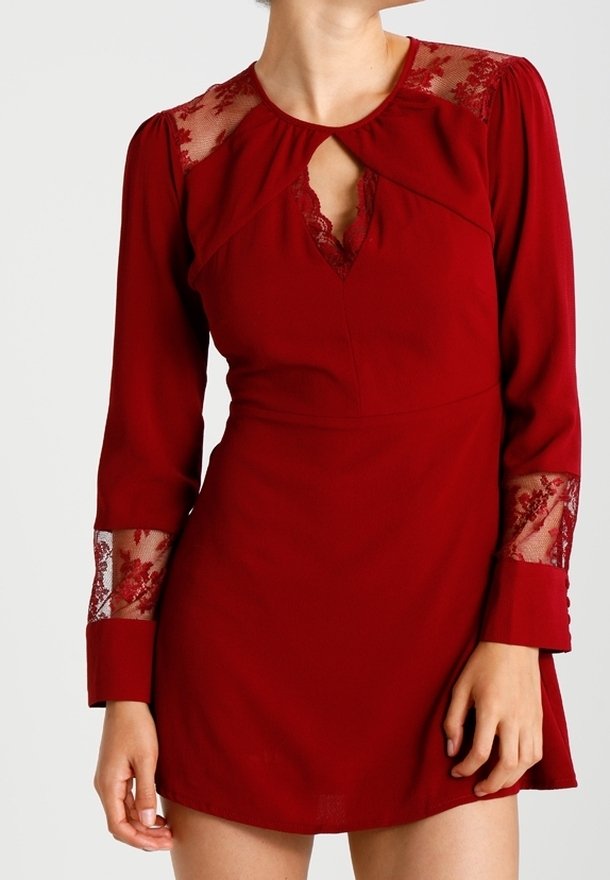 Červené šaty s dlouhými rukávy, krajkovými detaily na ramenou a manžetách, výstřihem ve tvaru klíčové dírky, přiléhavým korzetem a rozšířenou sukní. Hladká textilie.