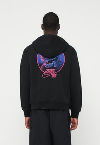 Osoba oblečená v čiernej mikine s kapucňou, na ktorej je farebná grafika hysčiaceho modrého tvora a ružový text "Nike SB" na zadnej strane.
