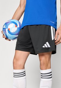 Μπλε αθλητικό πουκάμισο, μαύρο σορτς με λευκό λογότυπο Adidas, λευκές κάλτσες μέχρι το γόνατο με μαύρες ρίγες και πολύχρωμη μπάλα ποδοσφαίρου στο χέρι.