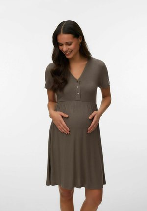 Kobieta w ciąży o długich ciemnych włosach, ubrana w brązową, krótkorękawową sukienkę na guziki, trzymająca się za brzuch i uśmiechająca się, stojąca na jednolitym tle.