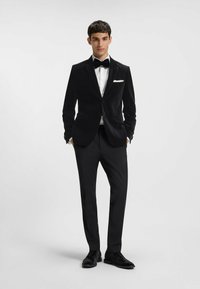 Zwarte fluwelen smokingjack met een sjalkraag, witte overhemden en een zwarte vlinderdas, gecombineerd met op maat gemaakte zwarte pantalon en schoenen.