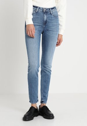 Schmal geschnittene hellblaue Jeans, getragen mit schwarzen Schnürschuhen und einem weißen Langarmshirt, stehend vor einem einfarbigen hellen Hintergrund.