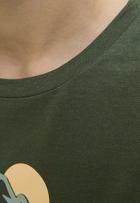 T-shirt en coton vert olive avec un col rond, une texture lisse et un design graphique détaillé en bleu clair et jaune sur le devant.