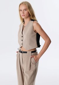 Beige cropped vest met een zwarte achterkant, gemaakt van een gestructureerde stof, met een v-halslijn en drie knopen. Gepaard met high-waisted beige broek.