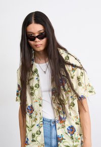 Kortærmet blomsterkravetrøje i lys gul med blå og orange mønstre, båret over en hvid tanktop, parret med denimshorts.