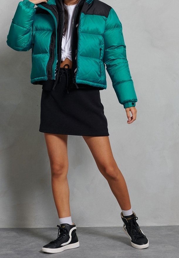 Personne portant une doudoune bleu sarcelle et noire, un top court blanc, une jupe noire, des chaussettes blanches et des baskets montantes noires, debout contre un mur gris.
