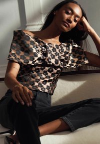Leoparda raksta tops ar nopietnu plecu siluetu, kas iekļauj strukturētu pūkainu piedurkni un vieglu audumu, kombinēts ar melnām džinsu biksēm.