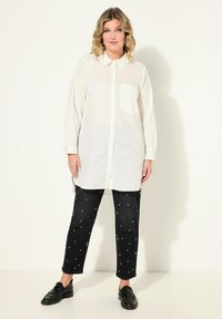 Hvid skjorte med knaplukning og brystlomme, parret med sorte jeans med hvide prikker. Sorte loafers fuldender outfittet.