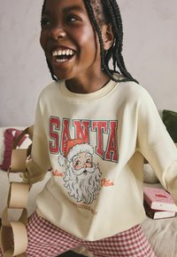 Kremi sweatshirt dengan cetakan "SANTA" merah dan wajah kartun Santa. Dipakai dengan celana kotak-kotak. Latar belakang mencakup perabotan lembut dalam nada netral.