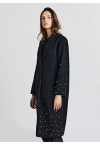 Cappotto di lana nero con maniche lunghe, caratterizzato da accenti in perle lungo le maniche e una chiusura con un solo bottone. Collo classico e vestibilità morbida.